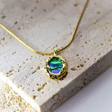 A gold chain necklace with a textured gold pendant featuring an iridescent green and blue abalone shell inlay, displayed on a light beige stone tile.