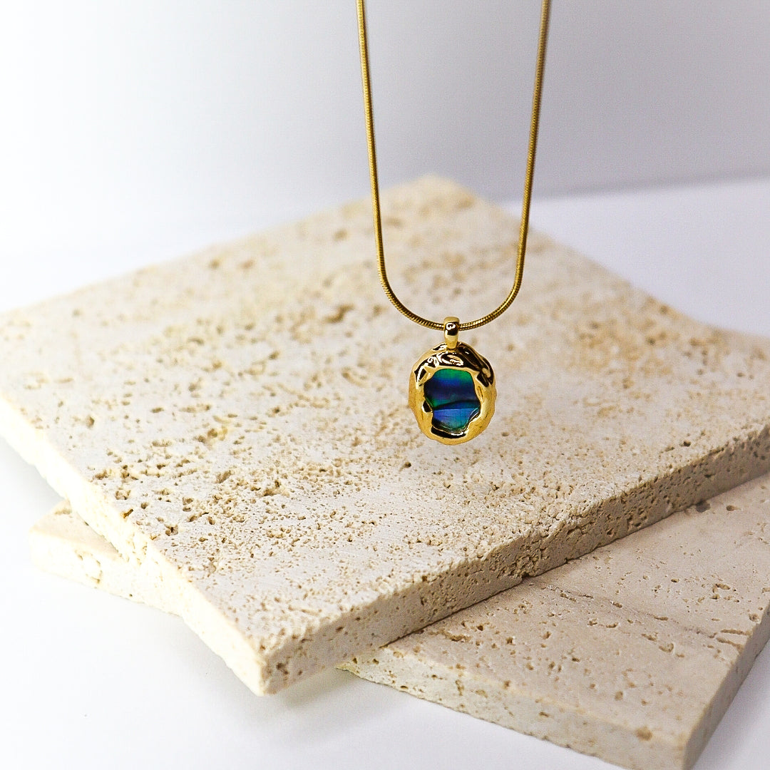 A gold pendant necklace with a textured, organic-shaped bezel holding a deep blue iridescent stone, displayed on a stack of light beige travertine tiles against a white background.
