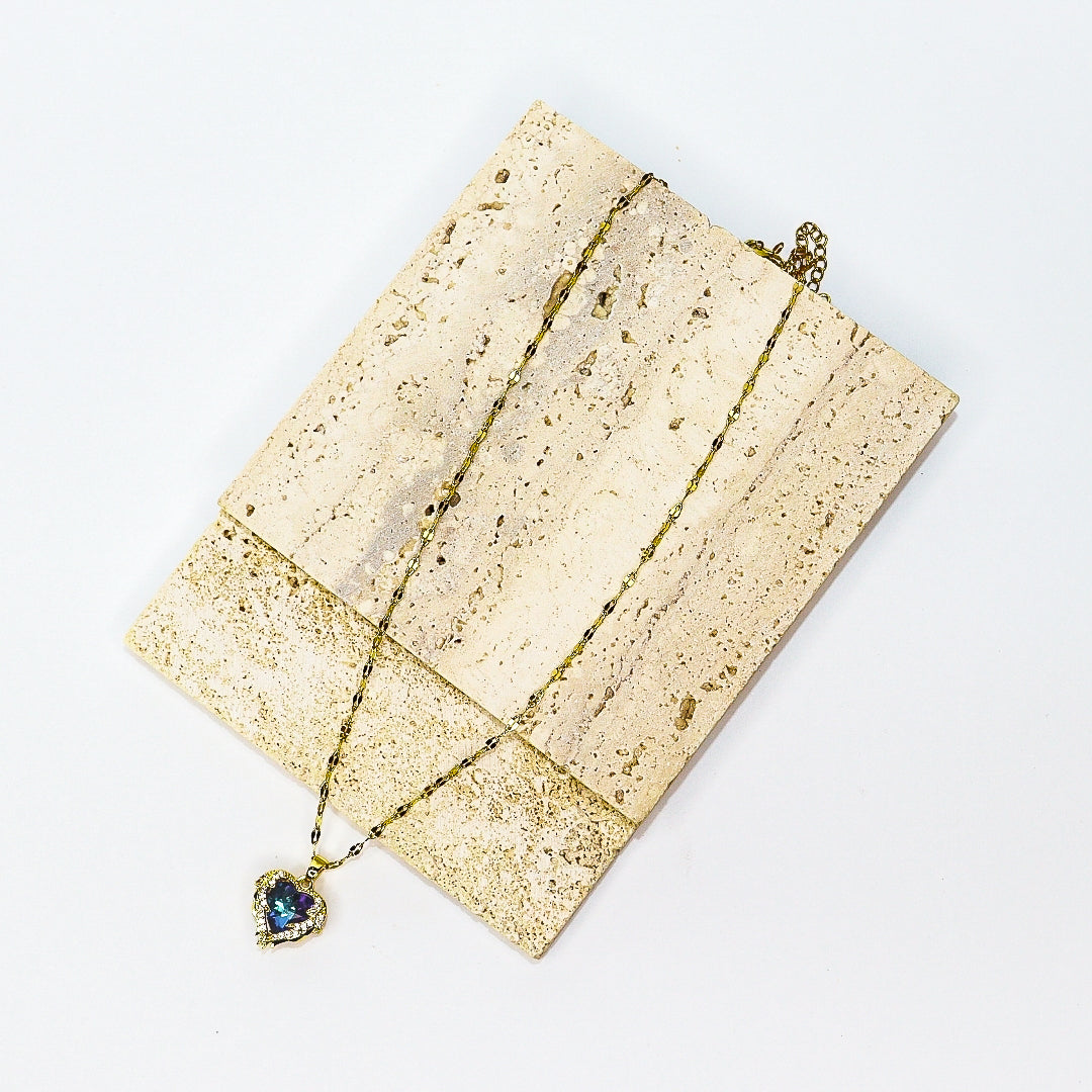 A gold chain necklace with a heart-shaped pendant featuring a blue iridescent stone, displayed on a textured beige stone block against a white background.