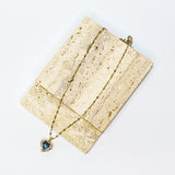 A gold chain necklace with a heart-shaped pendant featuring a blue iridescent stone, displayed on a textured beige stone block against a white background.