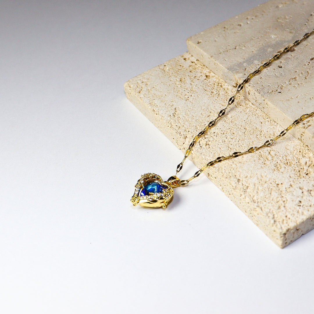 A gold chain necklace with a heart-shaped pendant featuring a blue gemstone, styled on a textured beige stone block against a white background.