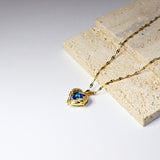 A gold chain necklace with a heart-shaped pendant featuring a blue gemstone, styled on a textured beige stone block against a white background.