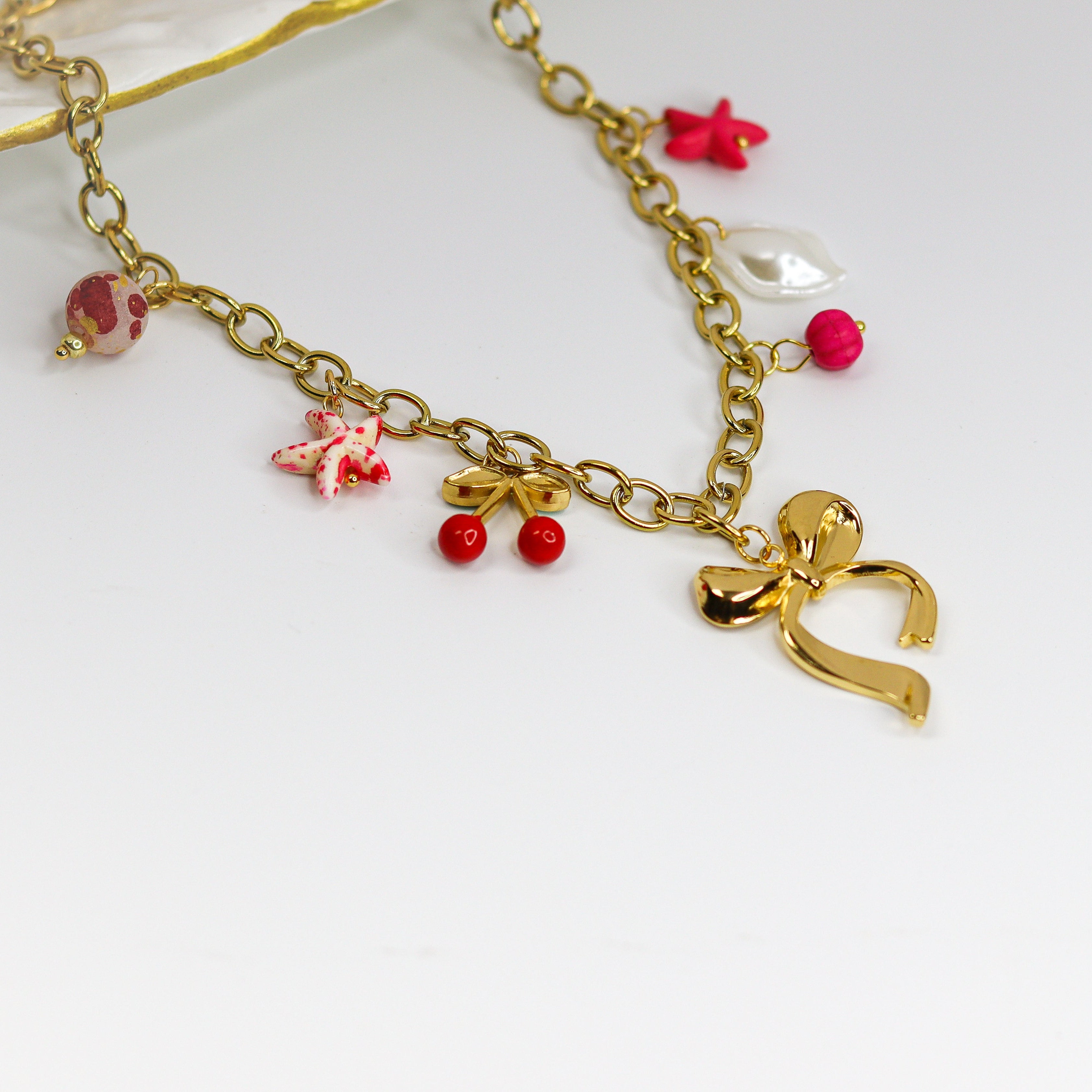 Chunky gold-tone chain necklace with statement bow and colorful enamel charms — cherries, starfish, beads, and a pearl drop.