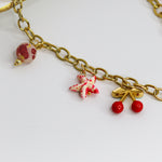 Chunky gold-tone chain necklace with statement bow and colorful enamel charms — cherries, starfish, beads, and a pearl drop.