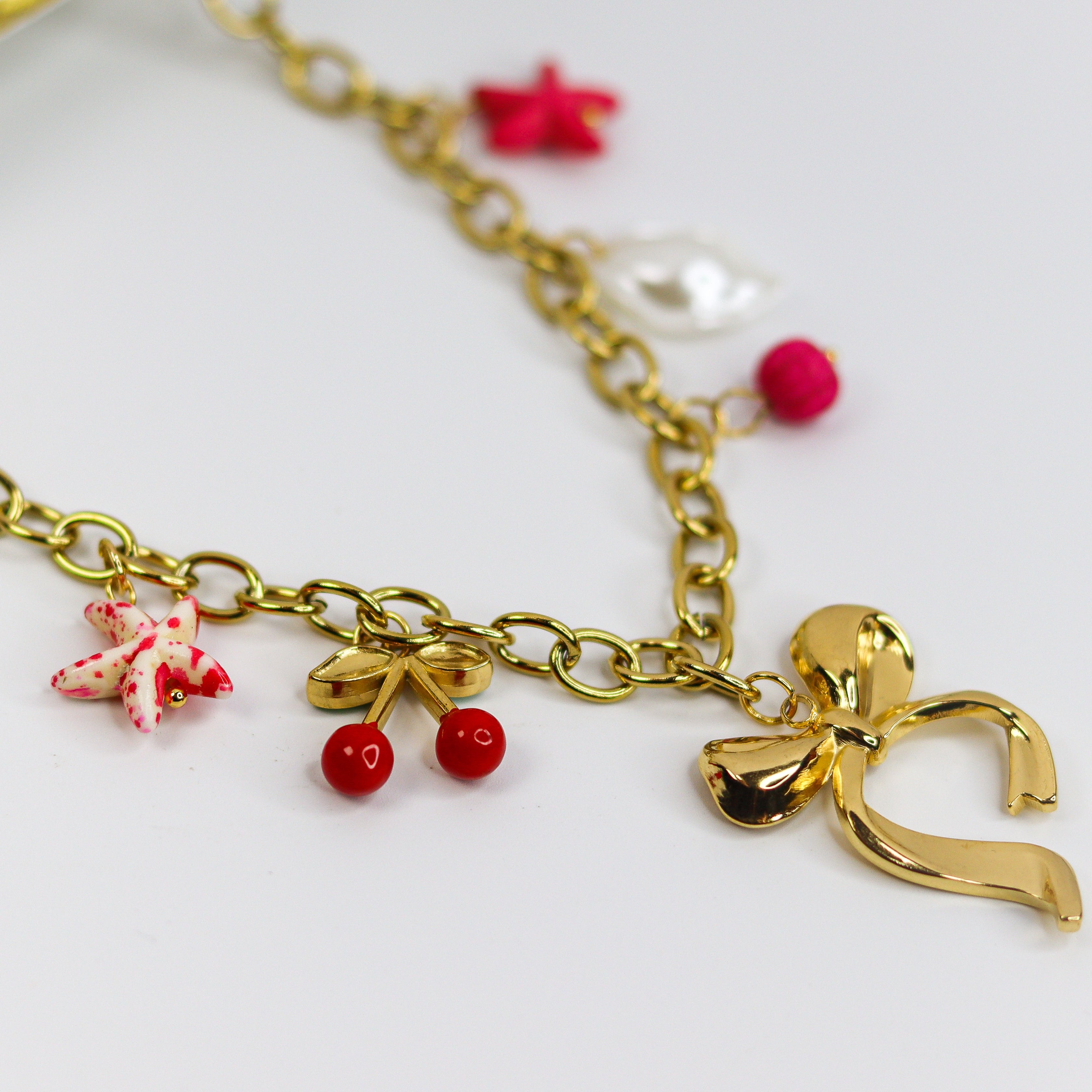 Chunky gold-tone chain necklace with statement bow and colorful enamel charms — cherries, starfish, beads, and a pearl drop.