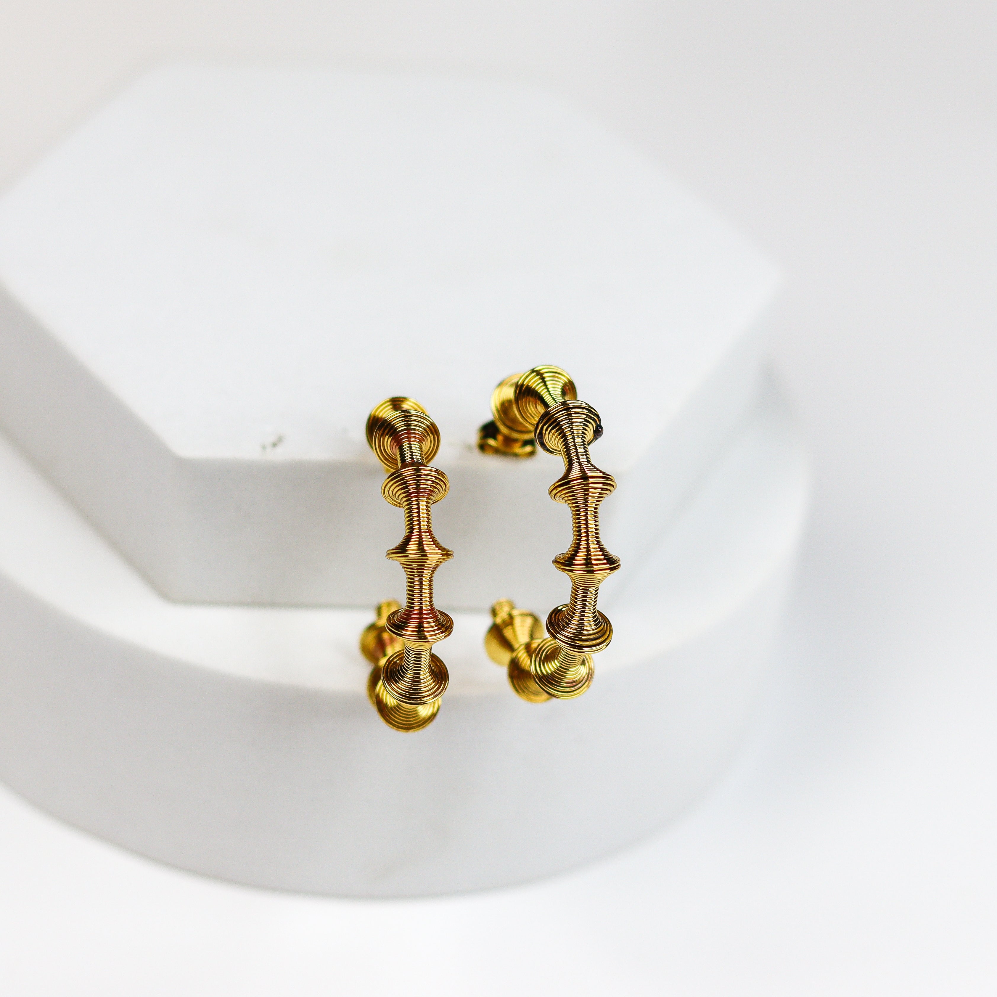 A pair of gold hoop earrings with a textured, organic leaf-like pattern, displayed on a white hexagonal jewelry stand against a clean white background.